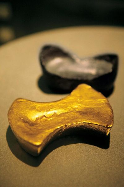 图片中近景是一块束腰形的明代50两金锭，远景是同时代的银锭，都藏于中国钱币博物馆。除了束腰形，金锭还有方形的“方锭”、圆形的“圆锭”、锤子形的“锤形锭”、马鞍形的“马鞍锭”，这些都是根据锭的形状所取的形象名字。不过明代黄金已不是主要货币，更多的是财富的象征。